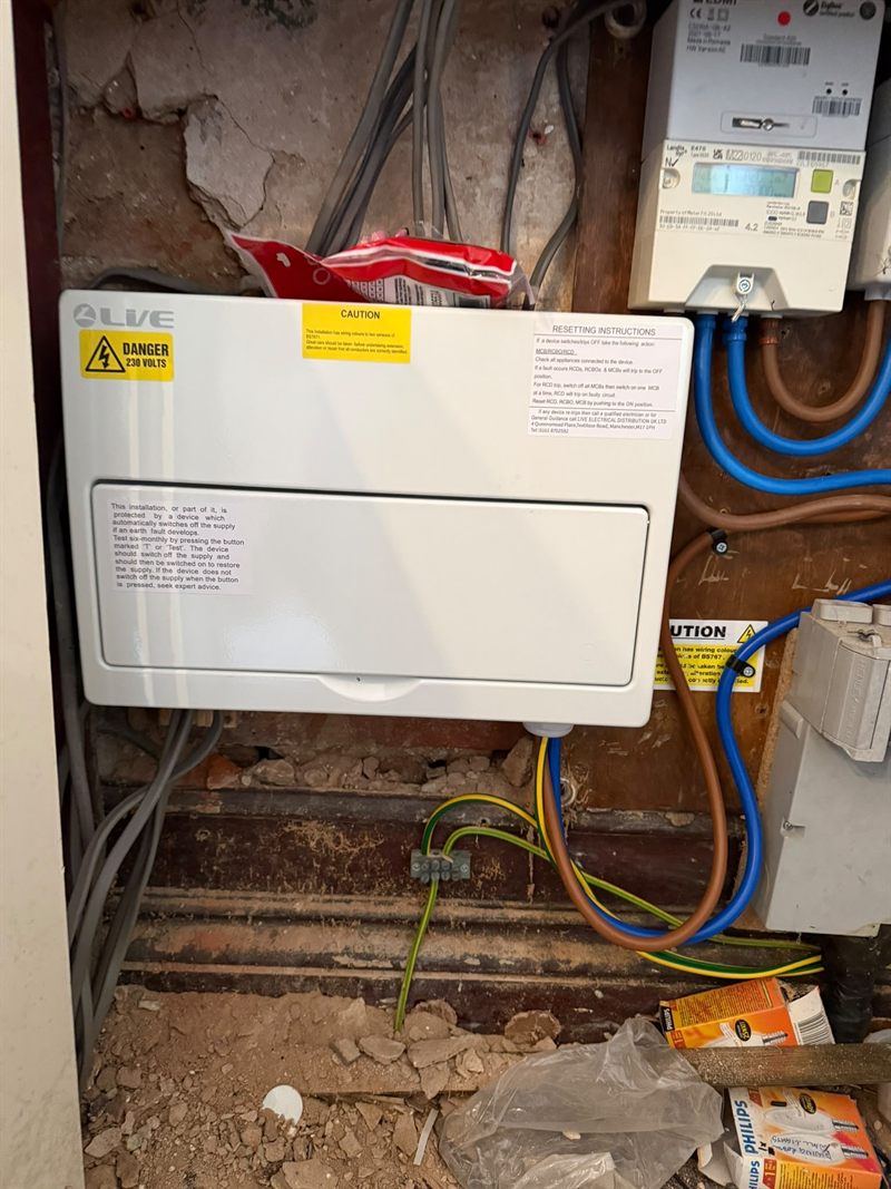 The same consumer unit closed, sitting beside the electricity meter with the tails run in and the earthing block below.