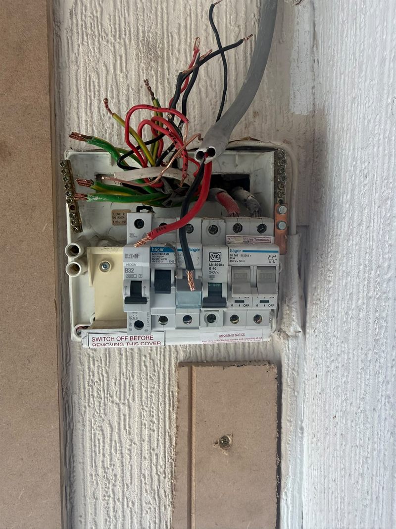 The board before replacement: an ageing plastic consumer unit with the cover removed, wired in the old red and black colours, with mixed makes of breaker fitted over the years.